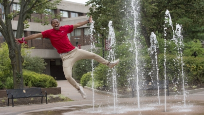 Male student jumps in the air in front a fountain sprsying water. He's wearing khaki pants and red Rutgers shirt.