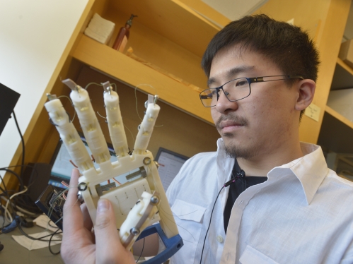 Young Asian male student wearing eyeglasses working with robotic hand.