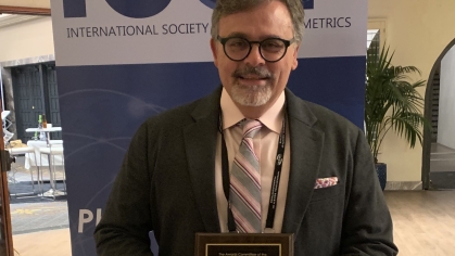 Man with gray hair and glasses wearing a suit and tie, poses with an award plaque in front of a banner that says ISOP.