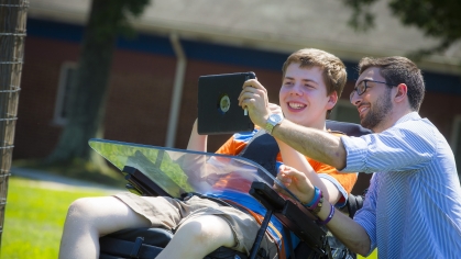 Male college student engages with young boy who is in a wheelchair. They are smiling and looking at an iPad.