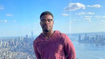 Black male with eyeglasses standing in the foreground of a cityscape. 