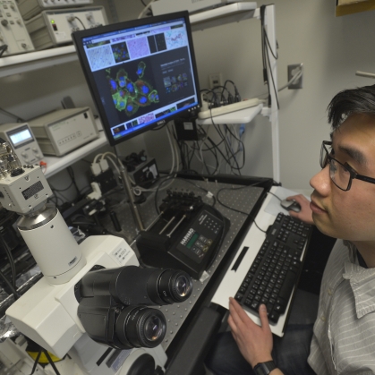 Male student with black hair and glasses looks at computer monitor alongside a confocal microscope.