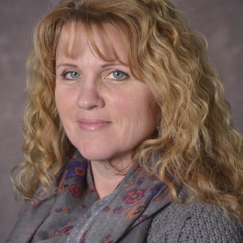 head shot of woman with long blonde curly hair wearing a grey sweater with a grey scarf draped aroung her neckaround