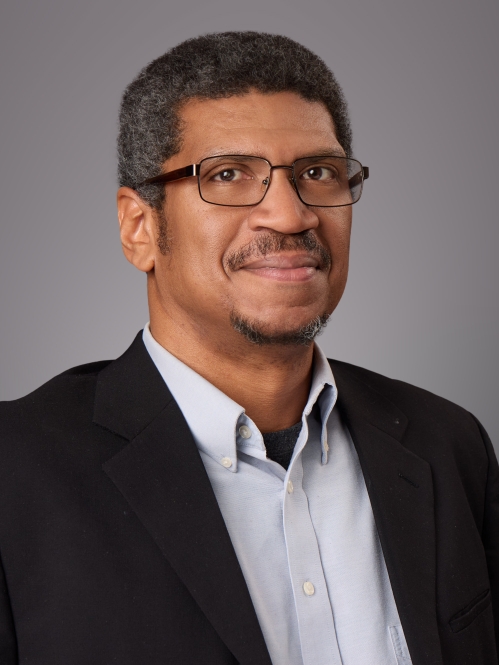 Headshot of Black male with eyeglasses wearing a light grey button down shirt and a dark suit jacket.