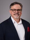 Headshot of male with short hair, grey beard and moustache, and eyeglasses wearing a black suit jacket, white button down shirt, and a red lapel handkerchief.