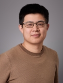 Headshot of male with short black hair and eyeglasses wearing a brown shirt.