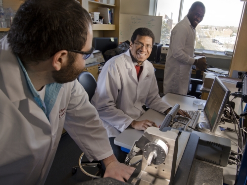 Male black professor with two male gradaute students wearing white lab coats conducting research in laboratory 