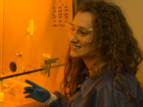 female graduate student with long curly brown hair working in research laboratory wearing safety goggles, blue lab coat, and blue latex gloves