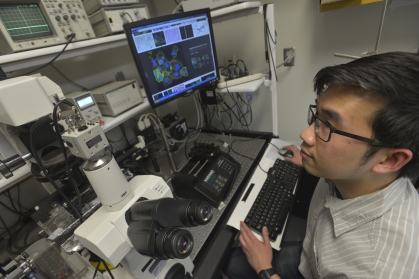 Male student with black hair and glasses looks at computer monitor alongside a confocal microscope.