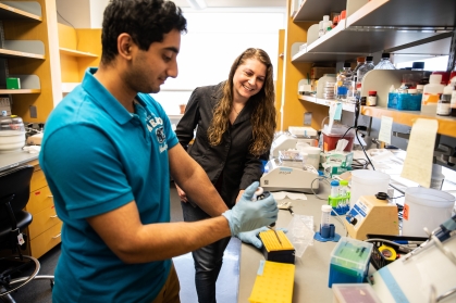 Female professor in a science lab instructing a male student who is using a syringe to test material.