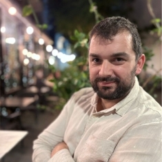 Headshot of male with short brown hair and beard with his arms crossed wearing a light cream colored button down shirt.